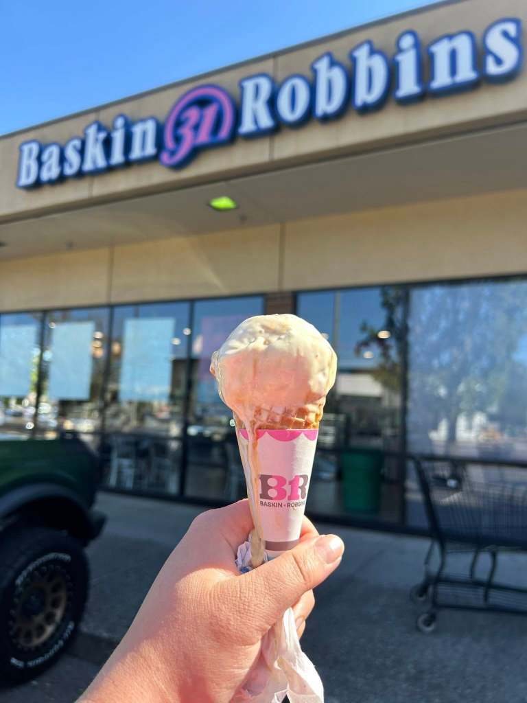 Someone holding a scoop of ice cream on a cone in front of Baskin-Robbins.